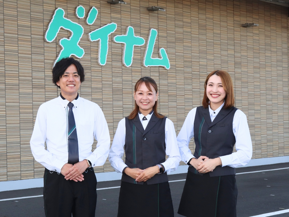 株式会社ダイナム 社名の前で笑顔で並ぶ３人の社員